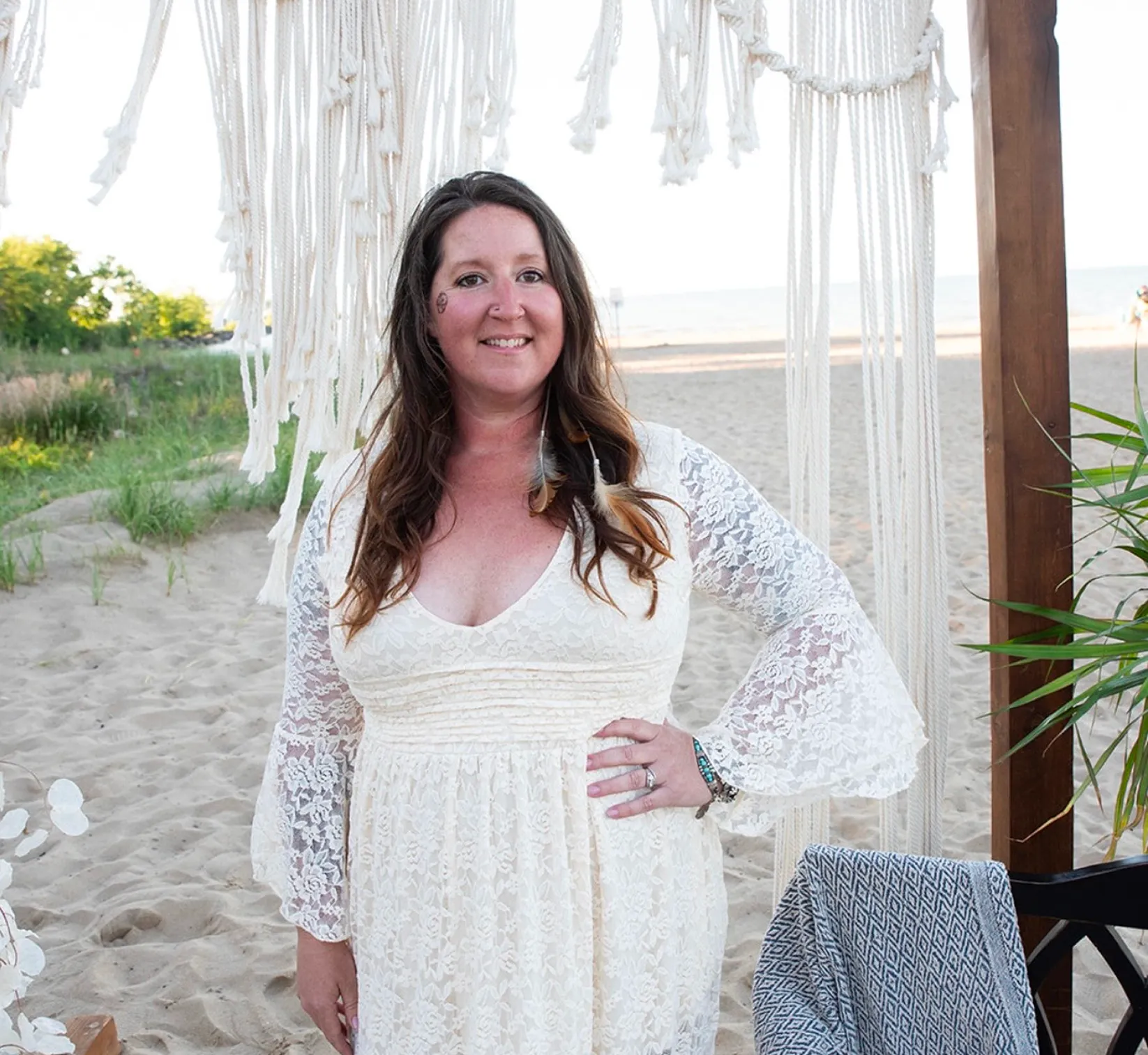 Woman in a dress standing on the beach, surrounded by a chair, a plant, a wooden pole